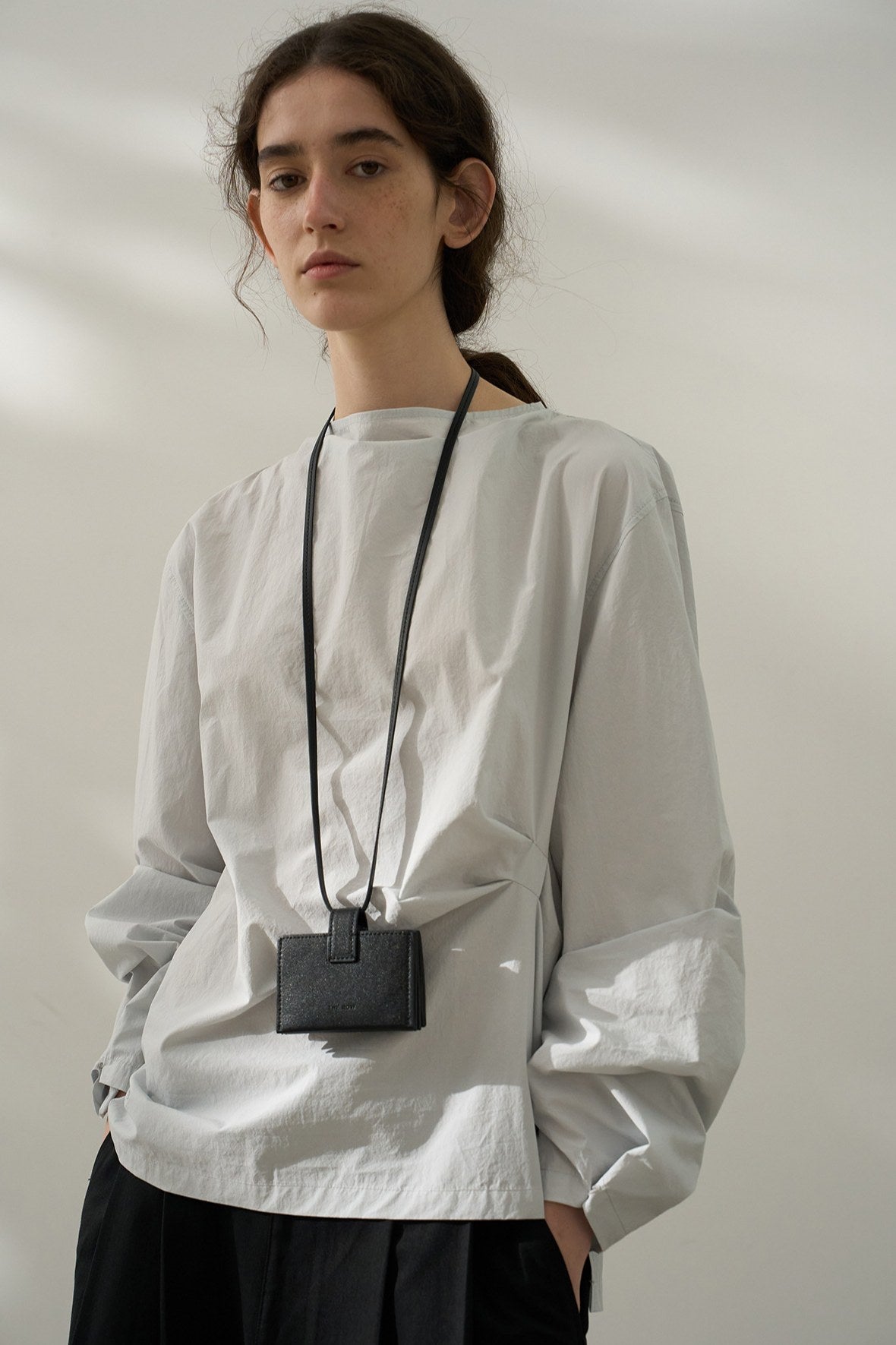 A woman modeling a long-sleeve grey blouse with a twisted waist design and a flowing collar, paired with black pants.